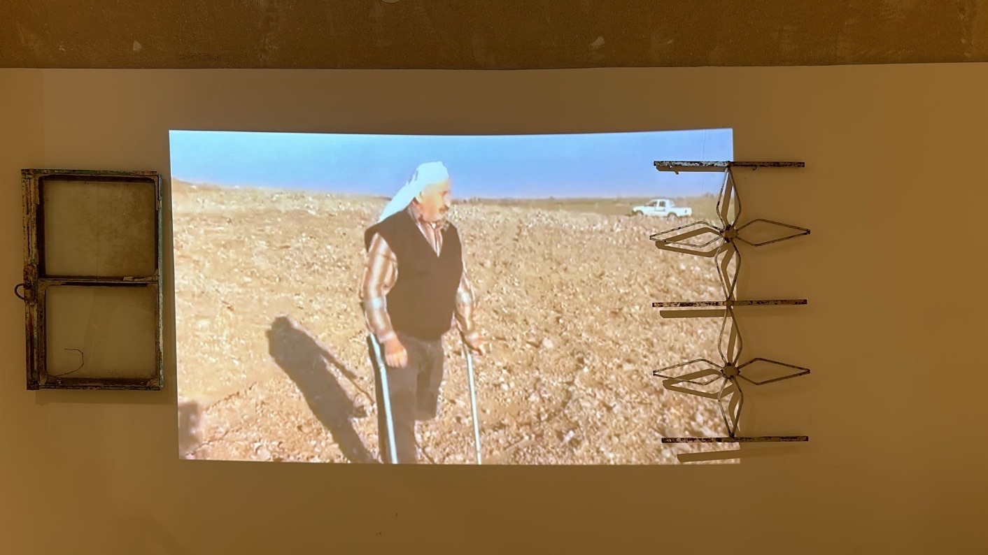 Video projection of an elderly man in a white headscarf, flanked by an old window and an iron window grille