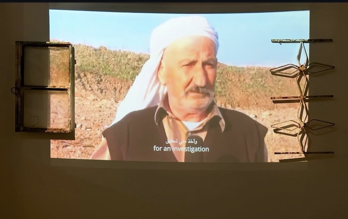 Projected video of a man with crutches standing in a ploughed field, between a window and an iron grille